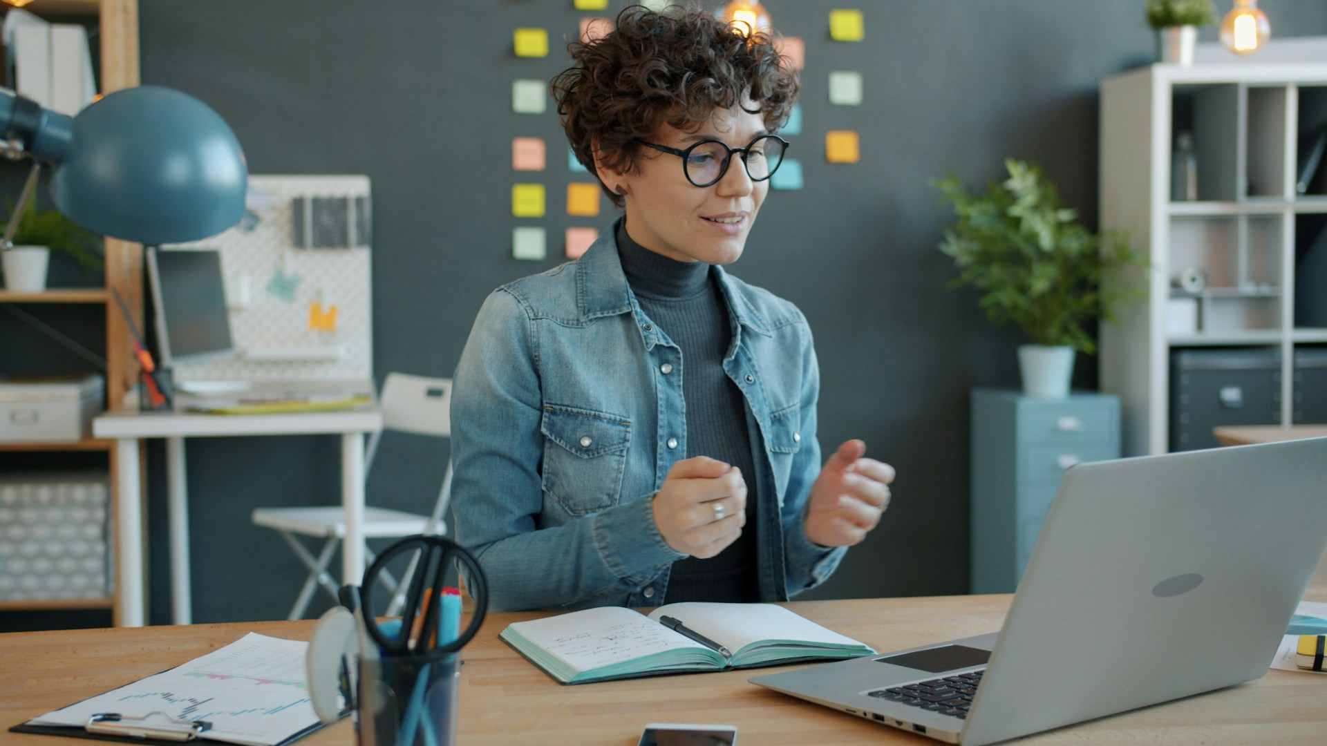 Professional on a video call at a desk with a notebook, charts, and a laptop